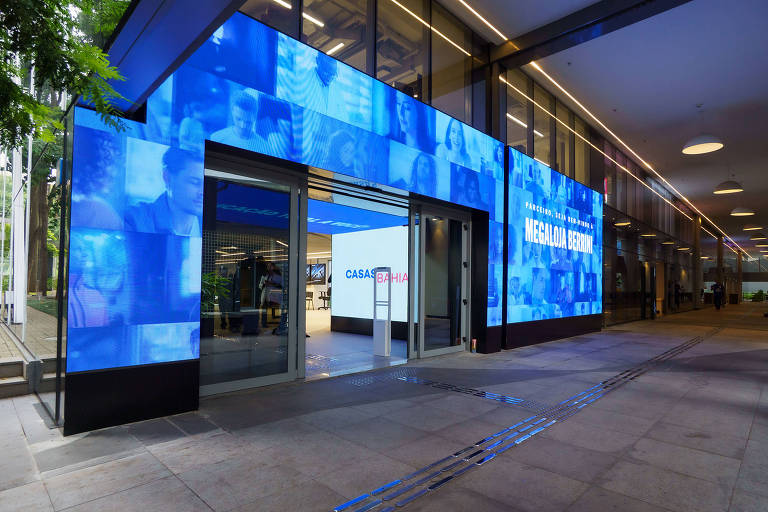 A imagem mostra a entrada da Casa Bahia, com uma fachada moderna e iluminada. As paredes externas são cobertas por telas digitais que exibem imagens em tons de azul. A porta de entrada é ampla e está aberta, revelando o interior do espaço, que é iluminado e minimalista. Um letreiro na parede interna diz 'NEGÓCIO ABERTO'.