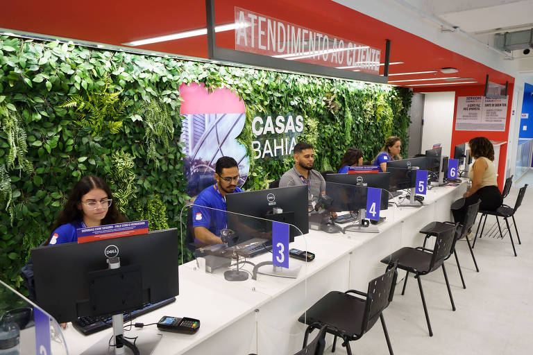 A imagem mostra um ambiente de atendimento ao cliente em uma loja. Há uma parede verde com plantas ao fundo e uma sinalização que diz 'ATENDIMENTO AO CLIENTE'. Vários atendentes estão sentados em mesas com computadores, e alguns deles estão usando camisetas azuis. No primeiro plano, há cadeiras pretas e mesas brancas. O ambiente parece moderno e bem iluminado.