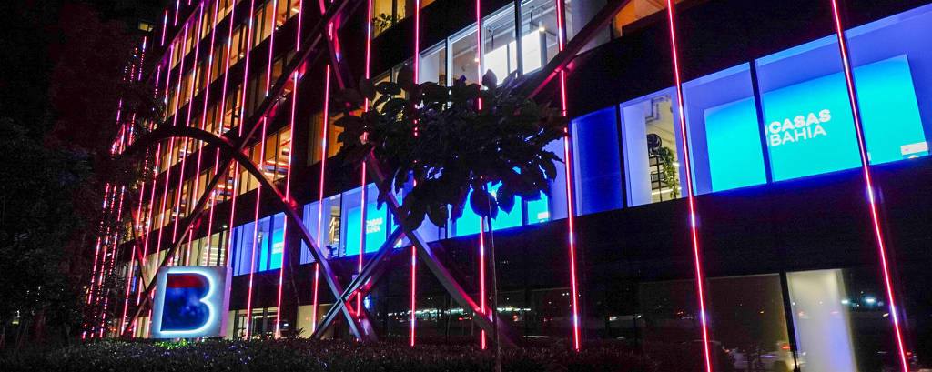 A imagem mostra a fachada de um edifício à noite, iluminada com luzes em tons de rosa e azul. À esquerda, há um grande letreiro em forma de 'B' e, à direita, janelas com iluminação azul que exibem o nome 'CASAS BAHIA'. A vegetação na frente é visível, com uma árvore pequena.