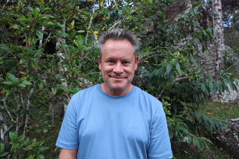 Um homem sorridente está posando em um ambiente natural, cercado por árvores e vegetação. Ele usa uma camiseta azul e está em pé, com um fundo de folhagens verdes e algumas estruturas rochosas ao fundo.