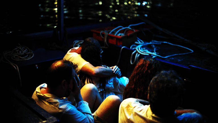 A imagem mostra um grupo de pessoas sentadas em um barco à noite. Algumas delas estão abraçadas, enquanto outras parecem pensativas. O ambiente é escuro, com reflexos de luz na água ao fundo. Há cordas e um recipiente vermelho visíveis no barco.