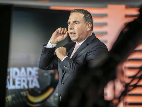 EXCLUSIVO F5 - Reinaldo Gottino posa para o F5 durante comando do Cidade Alerta, da Record *** Local Caption *** Um apresentador de televisão está em um estúdio, gesticulando com as mãos enquanto fala. Ele usa um terno escuro e uma gravata rosa. Ao fundo, é possível ver o nome do programa 'CIDADE ALERTA' em letras grandes. O ambiente é bem iluminado, com um design moderno.