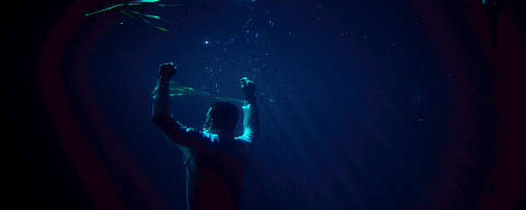 Cena da série Bateau Mouche: O Naufrágio da Justiça *** Local Caption *** A imagem mostra um homem submerso em água, com os braços levantados e os punhos cerrados. A luz que entra na água cria um efeito de brilho e reflexos, enquanto bolhas de ar sobem à superfície. Há também algumas plantas aquáticas visíveis na parte superior da imagem, e a cena é predominantemente iluminada por tons de azul e verde.