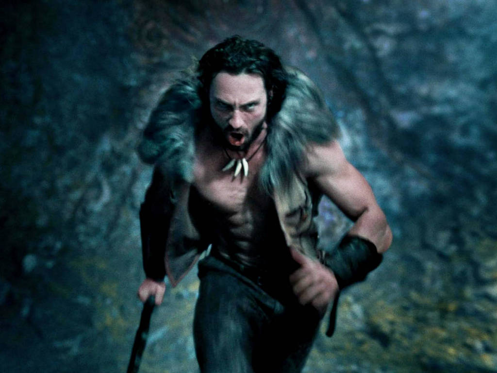 Cena do filme Kraven, o Caçador com Aaron Taylor-Johnson *** Local Caption *** Um homem musculoso e sem camisa está correndo em um ambiente escuro e rochoso. Ele usa uma roupa feita de pelagem e tem um colar de dentes. Sua expressão é intensa e ele parece estar gritando ou emitindo um som de batalha. O fundo é desfocado, com tons de azul e verde, sugerindo um cenário natural.