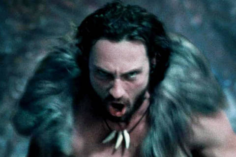 Cena do filme Kraven, o Caçador com Aaron Taylor-Johnson *** Local Caption *** Um homem musculoso e sem camisa está correndo em um ambiente escuro e rochoso. Ele usa uma roupa feita de pelagem e tem um colar de dentes. Sua expressão é intensa e ele parece estar gritando ou emitindo um som de batalha. O fundo é desfocado, com tons de azul e verde, sugerindo um cenário natural.