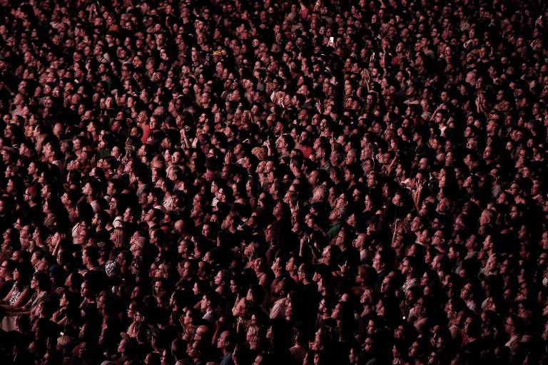 A imagem mostra uma grande multidão de pessoas reunidas, com rostos visíveis em meio a uma iluminação predominantemente escura e avermelhada. As pessoas parecem estar animadas, algumas levantando as mãos, enquanto outras estão concentradas na frente. A cena transmite uma sensação de energia e agitação.