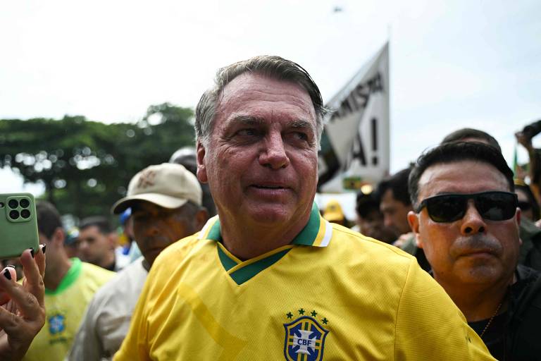 Um homem está usando uma camisa amarela com detalhes da seleção brasileira de futebol. Ele está cercado por outras pessoas em um ambiente ao ar livre, com árvores ao fundo. O homem parece estar sorrindo e é observado por outros indivíduos, alguns dos quais estão usando óculos escuros.