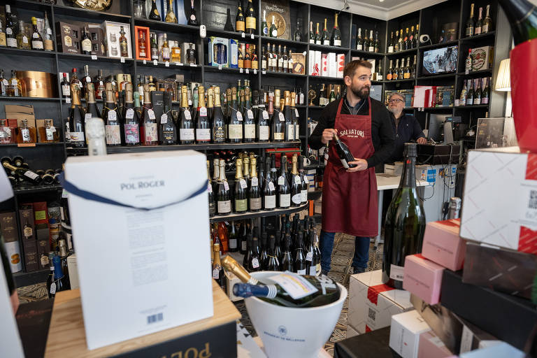 A imagem mostra um interior de uma loja de vinhos, com prateleiras cheias de garrafas de vinho. Um homem com um avental está em pé, segurando uma garrafa. Há caixas de vinho e outros produtos espalhados pelo chão.