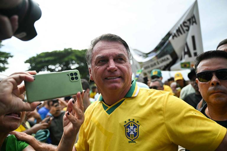 A imagem mostra um homem vestindo uma camisa amarela da seleção brasileira, cercado por uma multidão. Ele está sorrindo e acenando, enquanto uma pessoa à sua esquerda segura um celular. Ao fundo, há uma faixa com texto visível, e a cena parece ocorrer em um ambiente ao ar livre.