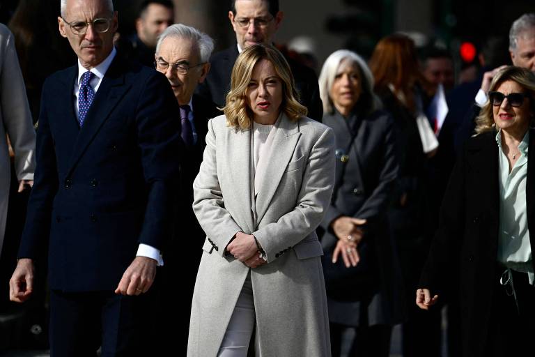 A imagem mostra um grupo de pessoas caminhando em um evento público. No centro, uma mulher com cabelo loiro e vestido claro parece estar em destaque. Ao redor dela, homens e mulheres vestindo trajes formais, incluindo ternos e casacos. O ambiente parece ser ao ar livre, com luz natural e um fundo desfocado.