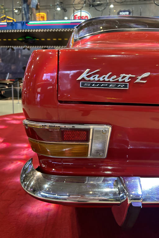 A imagem mostra a parte traseira de um carro vermelho modelo Kadett L Super. O emblema 'Kadett L Super' está visível na lateral do carro. A parte inferior do carro apresenta um para-choque cromado e as luzes traseiras são visíveis, com uma combinação de luzes vermelhas e âmbar. O fundo da imagem é desfocado, com um ambiente que parece ser uma exposição de automóveis.