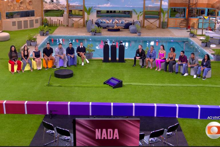 A imagem mostra um grupo de pessoas sentadas em um ambiente externo, com um gramado verde e uma piscina ao fundo. As pessoas estão organizadas em duas fileiras, algumas vestindo roupas coloridas. Na frente, há um palco com um painel que exibe a palavra 'NADA'. O ambiente parece ser parte de um reality show.