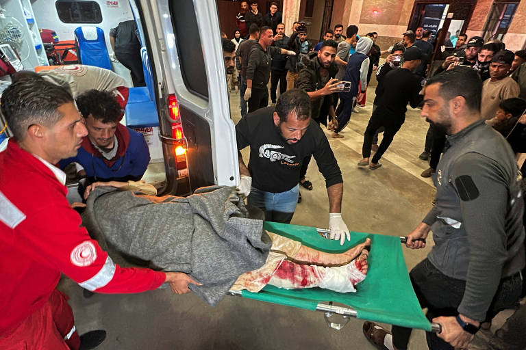 A imagem mostra uma equipe de socorristas transportando uma pessoa ferida em uma maca, coberta com um cobertor. A cena ocorre em um ambiente interno, com várias pessoas ao fundo observando. A maca é colocada em uma ambulância, e há evidências de sangue visíveis na roupa da pessoa ferida. Os socorristas estão concentrados na tarefa de ajudar a vítima.