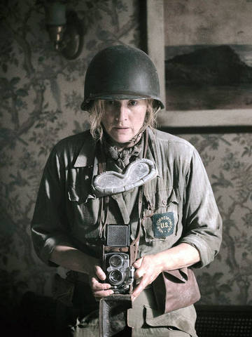 Cena do filme Lee com Kate Winslet *** Local Caption *** A imagem mostra uma mulher vestindo um uniforme militar e um capacete, segurando uma câmera fotográfica. Ela está em um ambiente interno, com uma parede decorada com papel de parede floral e uma pintura de paisagem ao fundo. A mulher tem uma expressão séria e parece estar focada. O uniforme possui um emblema que diz 'U.S.' e um acessório em forma de coração ao redor do pescoço.