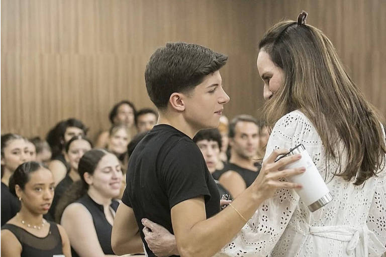 A imagem mostra um grupo de pessoas em um ambiente interno, possivelmente em uma sala de aula ou ensaio. No primeiro plano, um jovem de camiseta preta está conversando com uma mulher de cabelo longo e castanho, que usa uma blusa branca. Ao fundo, várias pessoas estão observando, algumas sorrindo. O ambiente é iluminado e as paredes são de madeira.