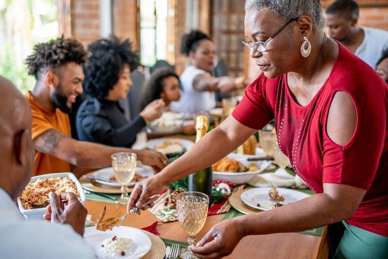 Uma mesa de jantar com várias pessoas reunidas, desfrutando de uma refeição. Uma mulher de cabelo curto e óculos está servindo comida a um homem careca que está sentado à mesa. Outros membros da família estão visíveis ao fundo, alguns se servindo e outros conversando. A mesa está decorada com pratos, copos e comida variada.