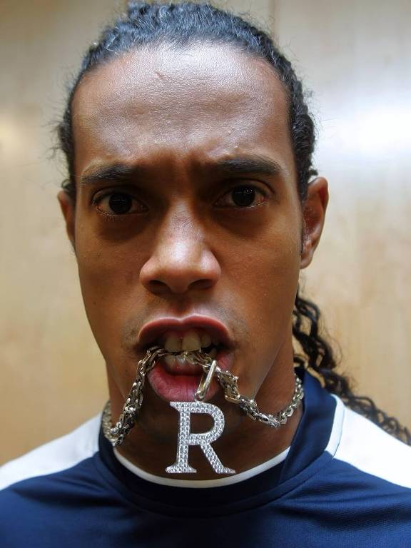 Um homem com cabelo longo e preso em um rabo de cavalo está posando para a foto. Ele tem uma expressão séria e está segurando uma corrente de colar com a letra 'R' na boca. O fundo é de madeira clara e ele está vestindo uma camiseta esportiva de cor escura com detalhes brancos.