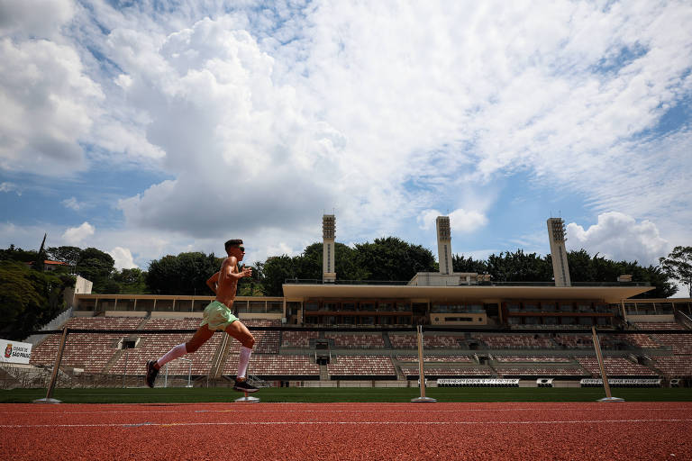 Um atleta está correndo em uma pista de atletismo, com um estádio ao fundo. O céu está parcialmente nublado, com nuvens brancas e um fundo azul. O estádio tem arquibancadas vazias e torres de iluminação visíveis.