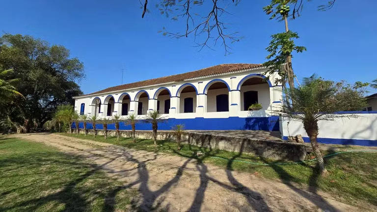 A imagem mostra uma casa de estilo colonial com arcos, pintada de branco e azul. A estrutura possui um telhado de duas águas e está cercada por um gramado verde. À frente, há um caminho de terra que leva até a casa, e sombras de árvores podem ser vistas no chão.