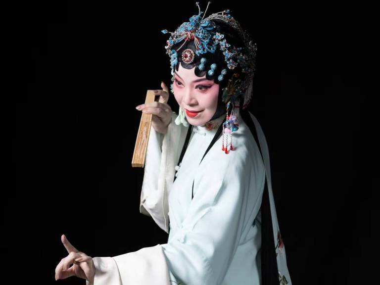 A imagem mostra uma artista de ópera chinesa vestida com um traje tradicional de cor azul claro， adornado com bordados florais. Ela está segurando um objeto em uma das mãos e fazendo um gesto expressivo com a outra. Seu cabelo está elaborado com enfeites coloridos e ela usa maquiagem característica da ópera， com destaque para os olhos e lábios. O fundo é escuro， destacando a figura da artista.