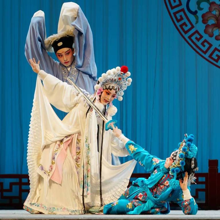 A imagem mostra três artistas em trajes tradicionais de ópera chinesa， em uma apresentação. O fundo é azul， com um padrão decorativo circular. Um dos artistas， à esquerda， está em uma pose dramática， enquanto outro， no centro， segura uma espada e está em uma posição de destaque. O terceiro artista， à direita， está ajoelhado， com um olhar expressivo. Os trajes são elaborados， com detalhes coloridos e acessórios típicos da ópera.