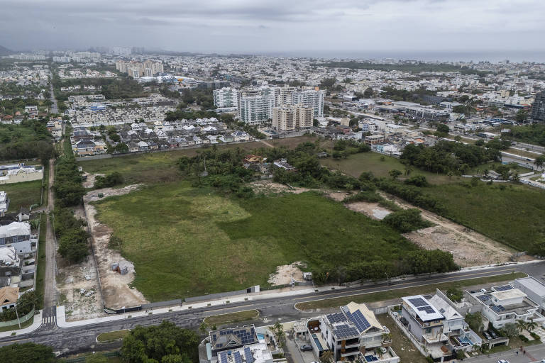 Imagem aérea de uma área urbana com terrenos vazios e construções. À esquerda, há uma estrada com casas e edifícios ao fundo. O céu está nublado, e a vegetação é visível em algumas partes do terreno. A cidade se estende ao fundo, com várias edificações e ruas.