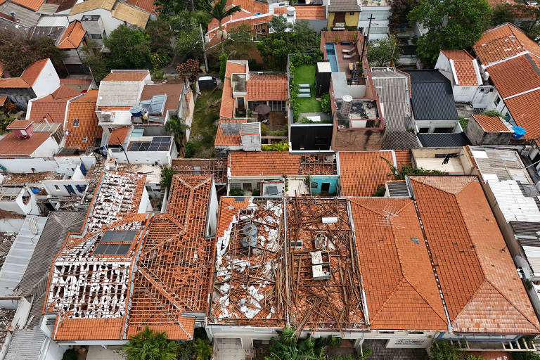 Imagem aérea mostrando uma área urbana com diversos telhados. Os telhados são predominantemente de cerâmica vermelha, com algumas áreas danificadas e expostas. Há uma mistura de casas e edifícios, com algumas áreas verdes visíveis entre as construções.