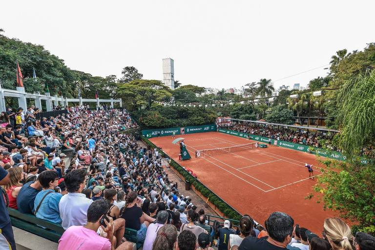 A imagem mostra um torneio de tênis em uma quadra de saibro, com uma grande quantidade de espectadores nas arquibancadas. O público está assistindo a uma partida, e a quadra está cercada por árvores e vegetação. Ao fundo, é possível ver edifícios da cidade.