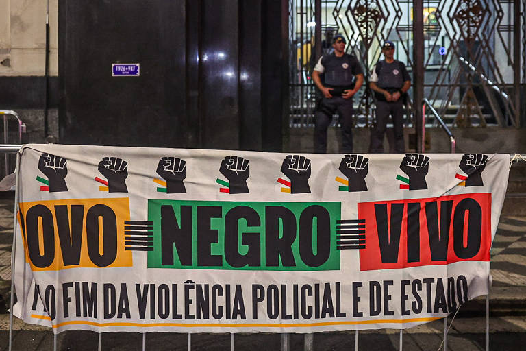 A imagem mostra uma faixa com as palavras 'POVO NEGRO VIVO' em letras grandes e coloridas, acompanhada de punhos cerrados. Abaixo, está escrito 'Pelo fim da violência policial e de Estado'. Ao fundo, há dois policiais em pé, vigiando a área. A faixa está presa a uma grade, e o ambiente parece ser urbano, com um edifício ao fundo.