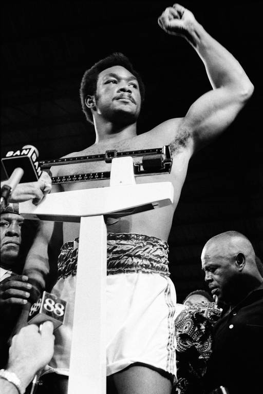 Um lutador de boxe está em uma balança, levantando o punho em sinal de vitória. Ele está sem camisa e usa calças de boxe com uma faixa na cintura. Ao fundo, há várias pessoas observando, incluindo um homem com uma expressão séria. O ambiente parece ser um evento de pesagem, com microfones e câmeras ao redor.