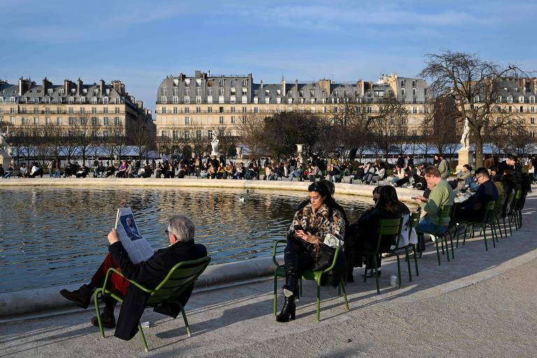 A imagem mostra várias pessoas sentadas em cadeiras verdes ao redor de um lago em um parque. Algumas pessoas estão lendo， enquanto outras usam seus celulares. Ao fundo， há edifícios com janelas e árvores. O céu está claro， indicando um dia ensolarado.