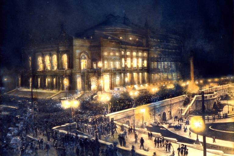 A imagem mostra um grande teatro iluminado à noite， com uma multidão reunida em frente. O edifício é imponente， com janelas grandes e detalhes arquitetônicos visíveis. A iluminação destaca a fachada do teatro， enquanto postes de luz ao redor iluminam a cena. A atmosfera é vibrante， com muitas pessoas na calçada e na rua.
