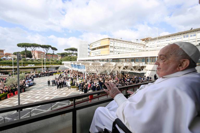 A imagem mostra o Papa sentado em uma cadeira de rodas, observando uma grande multidão reunida em um espaço aberto. Ao fundo, há um edifício moderno e árvores. O céu está parcialmente nublado.