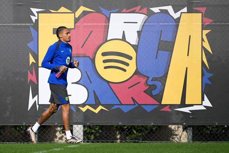 Um jogador de futebol está correndo em um campo de treino. Ele usa uma camisa azul e calças escuras. Ao fundo, há um mural colorido com a palavra 'FORÇA' em letras grandes e estilizadas, com elementos gráficos em várias cores.