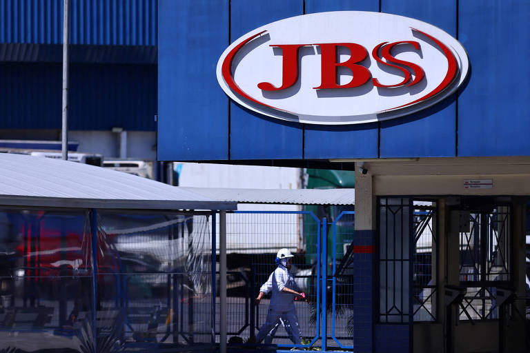 A imagem mostra a entrada de uma instalação da JBS, com um grande logotipo da empresa em destaque. Um trabalhador vestido com um uniforme branco e um capacete caminha em direção à entrada. Ao fundo, é possível ver caminhões estacionados e uma estrutura de metal.