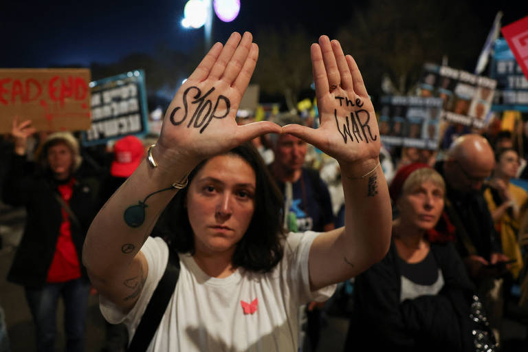 Uma mulher está em um protesto, levantando as mãos com as palavras 039;Stop039; e 039;the WAR039; escritas nelas. Ao fundo, há outras pessoas segurando cartazes e bandeiras, em um ambiente noturno.