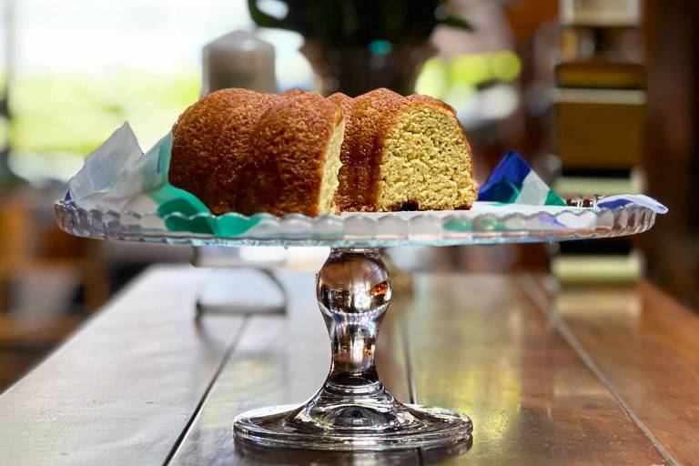 A imagem mostra um bolo em um prato de vidro sobre uma mesa de madeira. O bolo está cortado， revelando seu interior. Ao fundo， há um vaso com rosas vermelhas e uma decoração suave. A luz natural ilumina a cena， criando um ambiente acolhedor.