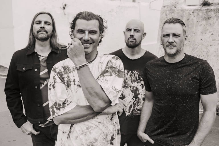Quatro homens posando em um ambiente urbano. O primeiro à esquerda usa uma jaqueta preta e tem cabelo longo. O segundo， no centro， está sorrindo e vestindo uma camiseta colorida. O terceiro， à direita， tem cabelo curto e usa uma camiseta preta. O fundo é composto por paredes brancas e edifícios ao fundo， com um céu nublado.