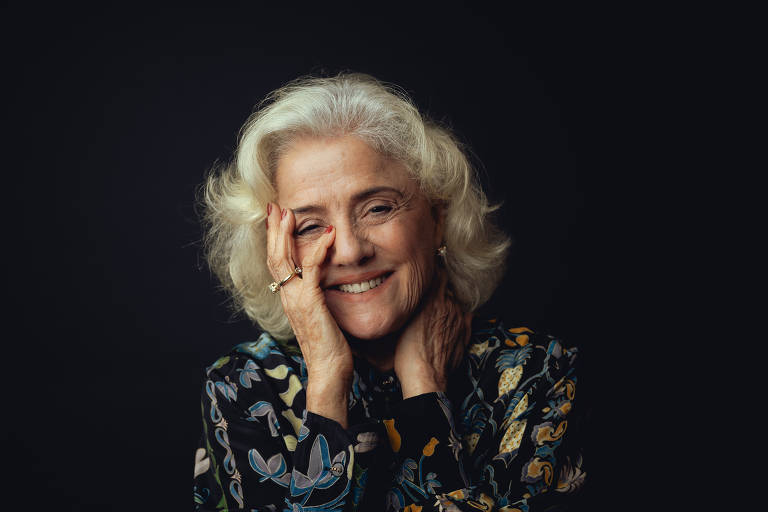 A imagem mostra uma mulher idosa com cabelo grisalho e ondulado, sorrindo suavemente. Ela está usando uma blusa escura com estampas coloridas de flores. A mulher está com uma das mãos no rosto, enquanto a outra mão segura levemente o pescoço. O fundo é escuro, destacando seu rosto e expressão amigável.