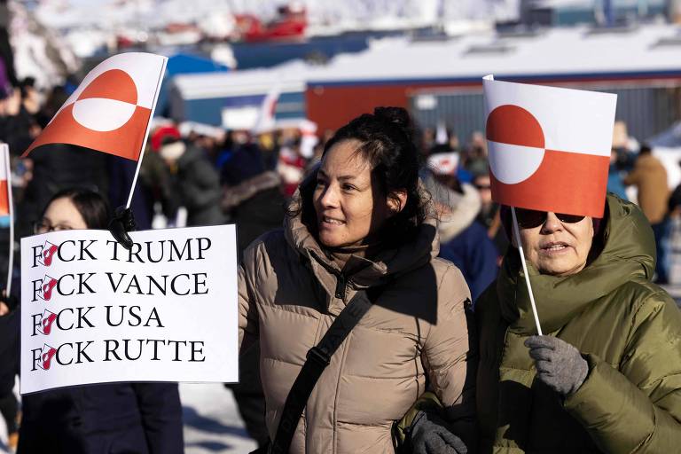 Duas mulheres participam de um protesto， segurando cartazes e bandeiras. Uma mulher à esquerda segura um cartaz com as palavras 039;FCK TRUMP， FCK VANCE， FCK USA， FCK RUTTE039;. A outra mulher à direita segura uma bandeira com um círculo vermelho e branco. O fundo mostra uma multidão e uma paisagem com neve.