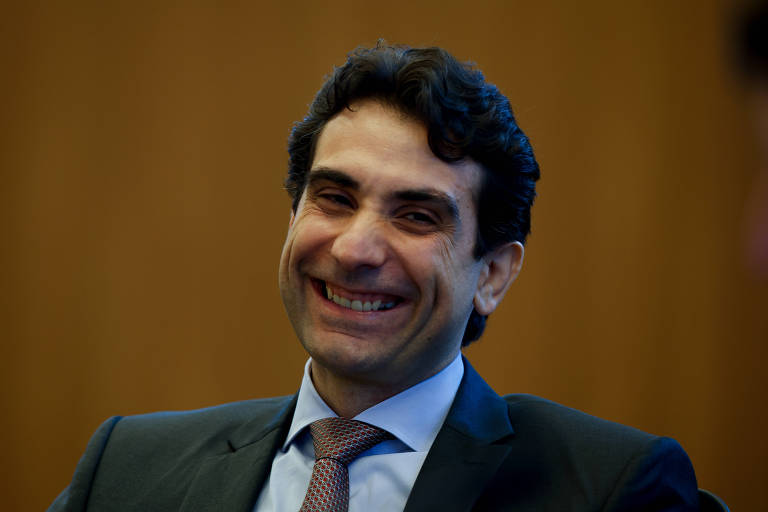 Um homem com cabelo escuro e ondulado, vestindo um terno escuro e uma gravata clara, está sorrindo. O fundo é de madeira clara, sugerindo um ambiente formal ou de negócios.
