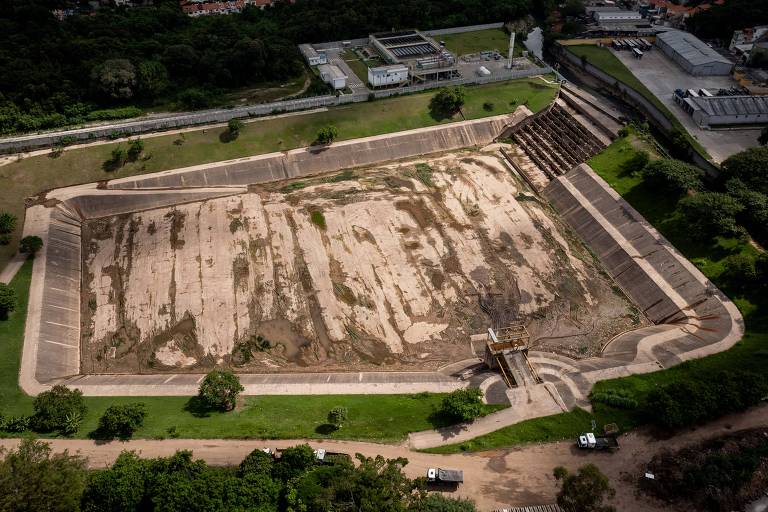 A imagem mostra uma vista aérea de um reservatório seco, com um grande espaço de areia exposta e paredes de contenção ao redor. No fundo, há vegetação e algumas construções. O reservatório parece estar em uma área urbana, com estradas e veículos visíveis nas proximidades.