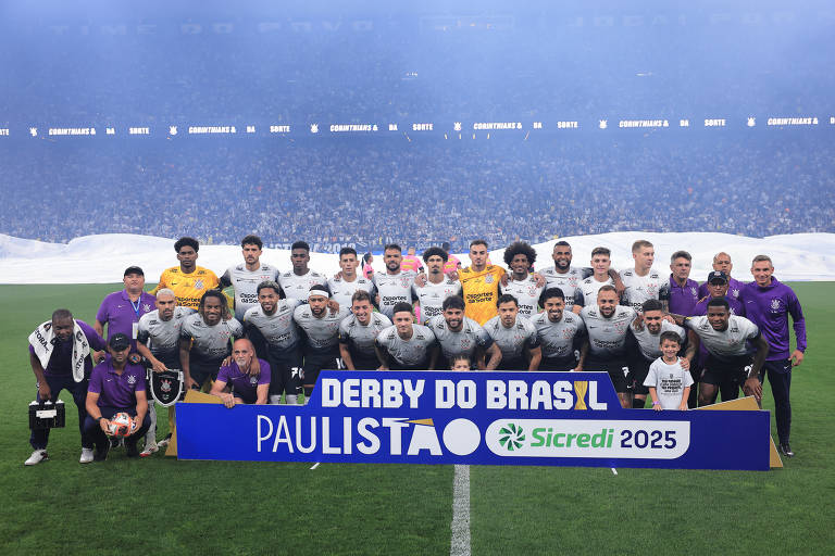 A imagem mostra um grupo de jogadores de futebol posando para uma foto em equipe. Eles estão em um campo de futebol, com um banner na frente que diz 'DERBY DO BRASIL PAULISTA 2025' e o logotipo da Sicredi. O time está vestido com uniformes de futebol, e há também alguns membros da comissão técnica e um troféu. O fundo é um estádio com uma grande quantidade de torcedores.