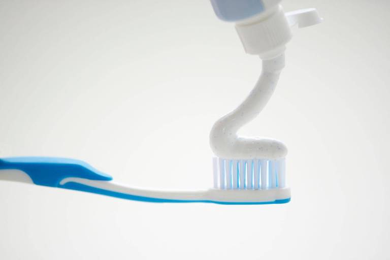 A imagem mostra uma escova de dentes azul e branca com creme dental branco sendo aplicado sobre as cerdas. O fundo é claro e desfocado.