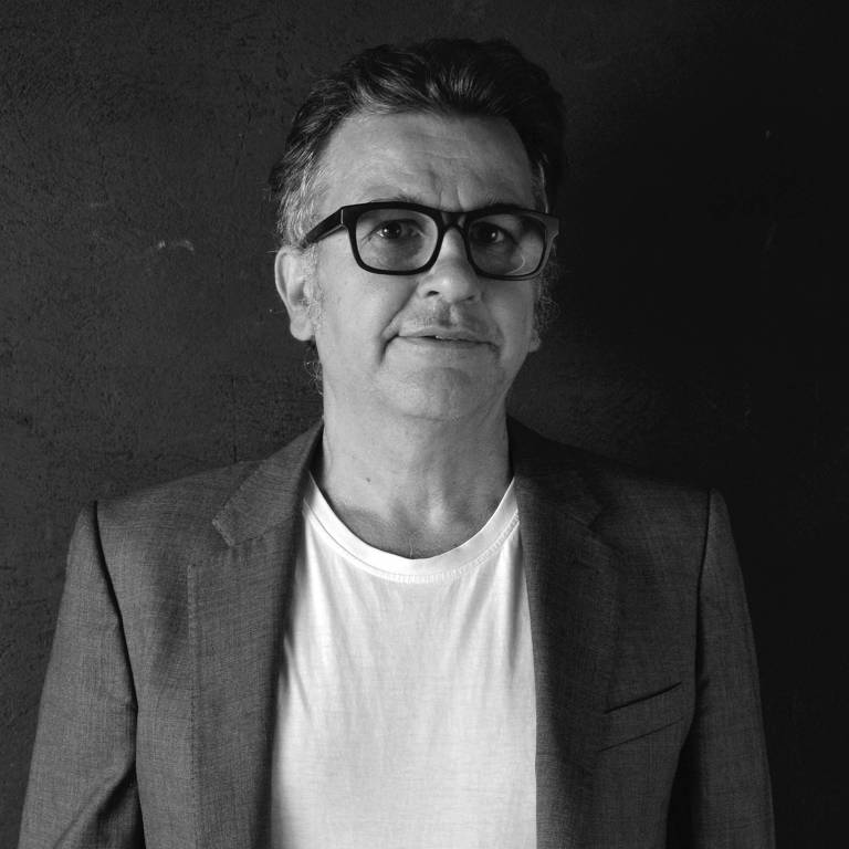 A imagem mostra um homem de meia-idade com cabelo grisalho e óculos, vestindo um blazer cinza sobre uma camiseta branca. Ele está posando em frente a um fundo escuro, com uma expressão neutra.