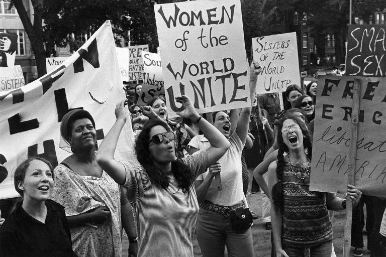 A imagem mostra um grupo de mulheres em um protesto, segurando cartazes com mensagens como 039;Women of the World Unite039; e 039;Sisters of the World Unite039;. As mulheres estão expressando suas opiniões com entusiasmo, algumas levantando os braços e outras gritando. O ambiente parece ser uma área pública, com árvores ao fundo.