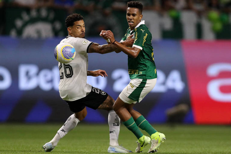 Dois jogadores de futebol estão em uma disputa pela bola em um campo. O jogador à esquerda， vestindo uma camisa preta com detalhes brancos e o número 26， está se afastando enquanto o jogador à direita， com uma camisa verde e detalhes brancos， tenta recuperar a posse da bola. Ao fundo， há uma torcida visível com bandeiras e um grande painel publicitário.