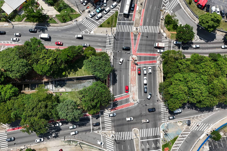 Imagem aérea de uma interseção movimentada, mostrando várias faixas de tráfego e semáforos. Há árvores ao redor da área, e diversos veículos, incluindo carros e ônibus, circulam pelas ruas. As faixas de pedestres são visíveis, assim como áreas de estacionamento.