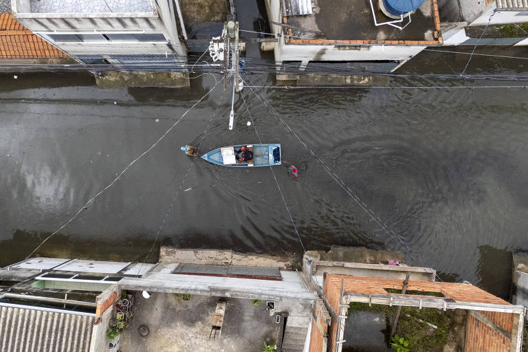 A imagem mostra uma vista aérea de uma rua inundada, onde uma pequena embarcação está navegando. As casas ao redor têm telhados de cerâmica e algumas áreas estão cobertas de água. Fios elétricos estão visíveis acima da água, e há objetos flutuando na superfície. A cena retrata um ambiente urbano afetado por enchentes.