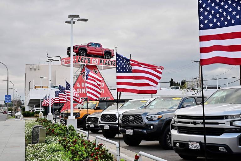 A imagem mostra uma concessionária de automóveis com várias bandeiras dos Estados Unidos dispostas ao longo da calçada. No fundo， há um grande letreiro com a frase 039;LET039;S GO PLACES039; e um carro vermelho em cima do letreiro. Vários veículos estão estacionados na frente， incluindo caminhonetes e SUVs. O céu está nublado.
